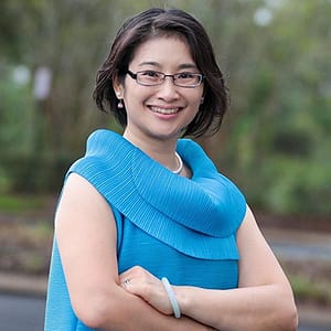 Su-I Hou wearing a blue blouse and smiling against a blurred nature background.