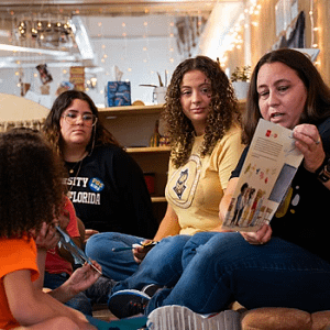 Three women from UCF’s School of Teacher Education sit on a classroom floor reading a book to young children listening in front of them.